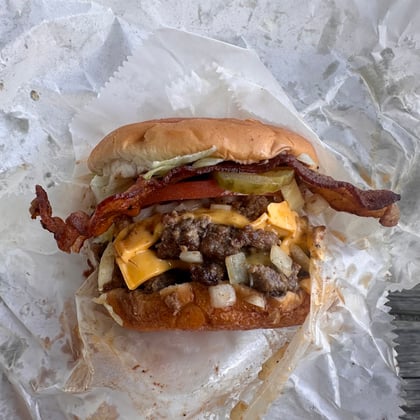 A bacon cheeseburger with pickles and onions on wax paper